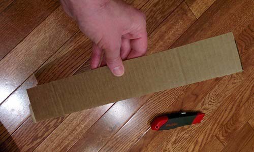 Hand holding small rectangle piece of cardboard over wood floor.